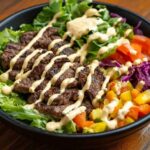 A colorful burger bowl with fresh ingredients and creamy sauce, placed on a rustic wooden table