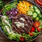 A colorful burger bowl with beef, lettuce, tomatoes, cucumbers, cheese, and dressing